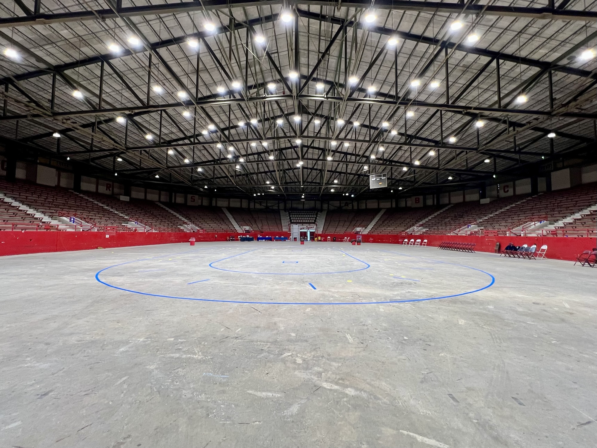 Wide-angle view of Blackham Coliseum in Lafayette, Louisiana, during setup for a roller derby bout. The concrete arena floor features a taped flat track outlined in blue, surrounded by red barriers and rows of empty chairs. The stands are empty, and the overhead lights brightly illuminate the expansive venue.